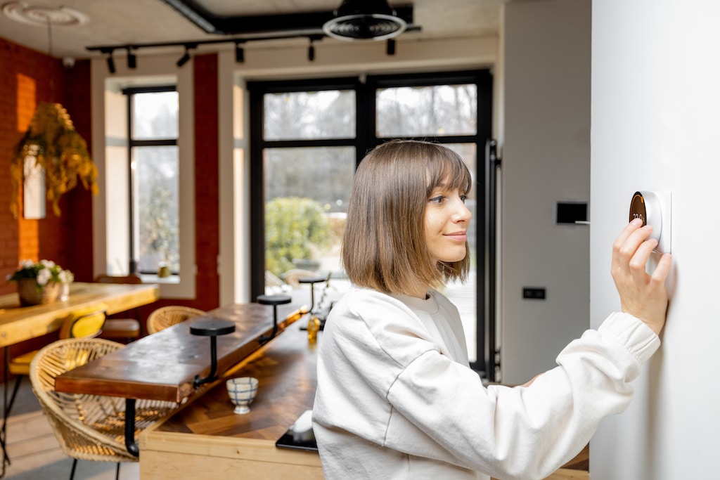 Woman controlling home temperature with electronic thermostat mounted on the wall in living room at home. Concept of modern technologies and smart home. Heating and AC repair