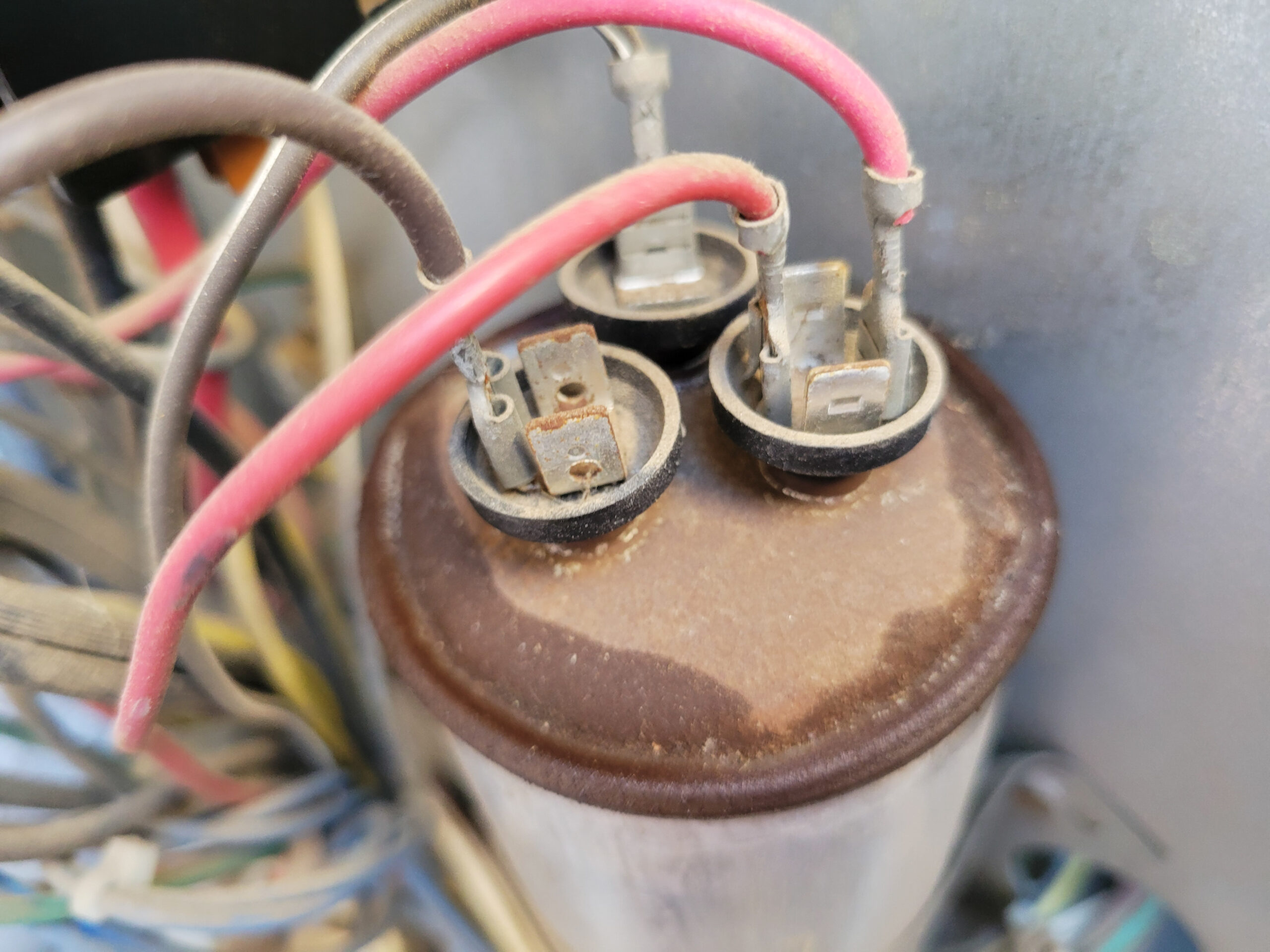 Rusty and leaking dual run capacitor terminals connected to red and brown wires in a dusty HVAC unit, Bad AC capacitor.